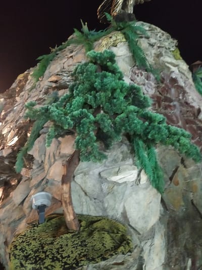The image shows a rocky outcrop covered with artificial plants, resembling a miniature mountain landscape. A small, dark brown trunk of a tree emerges from a mossy green surface at the bottom left. A white, disk-shaped light fixture is mounted on a gray arm extending from the rock face, also near the bottom left. The rock is composed of various shades of gray and brown, with some lighter colored stones. Lush, dark green artificial foliage, including pine-like branches and fern-like fronds, is strategically placed throughout the rock structure. The background is dark, suggesting an outdoor nighttime setting. There is no indication of the city of Sosnovka, Kyrgyzstan, in the image.