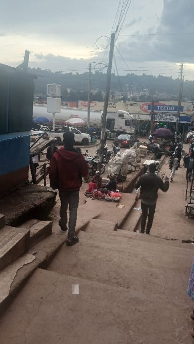 The image captures a dynamic street scene, likely in an urban or semi-urban environment, taken on an overcast day. The photo is oriented vertically, but when viewed horizontally (rotated 90 degrees clockwise), it shows a bustling street from a slightly elevated perspective.
In the immediate foreground on the right side, there are rough concrete or dirt steps leading down, with a portion of a person's legs and feet visible at the bottom right corner, suggesting the photo was taken from a raised vantage point. A small, white rectangular object rests on one of these steps.
The midground is dominated by the street and its activities. Several people are visible, including two main figures seen from behind in the central part of the image. One person wears a dark jacket and trousers, standing with arms slightly raised, appearing to be observing or interacting with something near the ground. Close by, another individual is seated on a colorful mat on the ground, possibly a street vendor. Further to the left and slightly closer to the viewer, a person in a red jacket and dark trousers stands, also facing away from the camera. The street is crowded with numerous motorcycles and scooters, some parked and others in motion, alongside a few cars or vans further down.
Buildings line both sides of the street, featuring various storefronts. A prominent power pole with a dense network of overhead wires runs across the upper midground against the sky. On one building facade in the mid-left, the brand name "TECNO" is clearly visible in white lettering on a blue background. Below it, another sign can be partially seen but is unreadable. On the left side of the street, a white, square-shaped structure, possibly a kiosk or small booth, is present.
The background reveals a hazy vista of distant buildings and what appear to be gentle hills under a cloudy sky, suggesting a broad urban or peri-urban landscape. The overall atmosphere is one of lively street activity, common in many busy city areas.