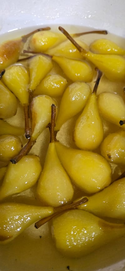 This media file is a close-up, top-down shot of many small, peeled pears that have been cooked and are submerged in a light-colored syrup. The pears are a vibrant yellow and have a smooth, glossy appearance. Some of the pears still have their stems attached, which are brown and twig-like. The syrup is clear and slightly viscous, with small bubbles visible in some areas, suggesting it is still warm. The overall impression is one of a sweet dessert or preserve. The scene is likely indoors, given the lighting. There are no visible people, text, or specific location cues to indicate the time of day, weather, or a precise setting beyond a kitchen or food preparation area. The pears themselves are the sole focus, highlighting their texture and color in the syrup.