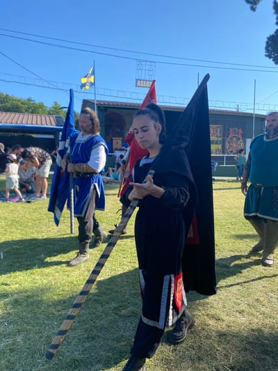 In Llanera, Spain, on a sunny day, a group of people in medieval-style attire are participating in an outdoor event. In the foreground, a young woman in a black hooded cloak with ornate trim is holding a black flag and a wooden staff. To her left, a man with long brown hair, dressed in a blue tunic and white shirt, carries a blue and yellow flag. Behind them, a man in a teal tunic is visible. The background shows a grassy field, tents, and spectators. The sky is clear and blue.