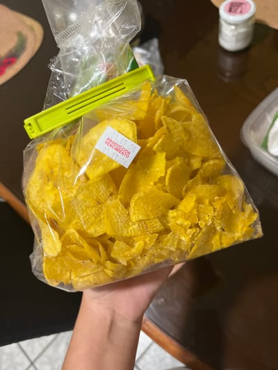 A close-up, eye-level shot features a hand holding a transparent plastic bag full of bright yellow, thinly sliced chips, likely plantain or banana chips. The chips vary in shape and size, displaying some darker specks, possibly seasoning or slight browning from frying. The bag is sealed at the top with a prominent neon green plastic clip.
Affixed to the front of the bag is a small white rectangular sticker with red text. The visible text reads: "21.05.25", followed by "PRODUCCION", and then "VENCIMIENTO 10.11.25", indicating production and expiration dates.
The background is mostly out of focus, revealing a dark, reflective surface that appears to be a table. To the left, a white bottle with a pink lid is partially visible, along with a clear plastic container and another plastic bag containing green, leafy contents, also secured with a similar green clip. On the right, a glimpse of a person's leg and part of a tiled floor suggests an indoor setting, such as a kitchen or dining area. The scene is illuminated by ambient indoor lighting. Given the context of San Luis, Peru, these chips are a typical local snack.