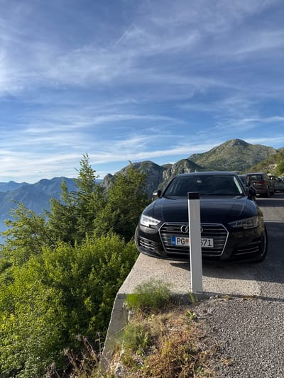 A black Audi A4 sedan is parked on the side of a winding mountain road with a panoramic view of mountains and a body of water. The car is in the foreground, facing towards the viewer, with its front bumper and license plate clearly visible. The license plate reads "PG 571". The road appears to be paved with asphalt and gravel. To the left of the car, lush green trees and foliage cascade down the mountainside, obscuring the lower half of the view. In the background, a range of rugged mountains stretches across the horizon, with a clear blue sky dotted with wispy clouds above. The scene suggests a scenic overlook in the mountainous region of Žanjev Do, Montenegro, likely a popular stopping point for travelers.