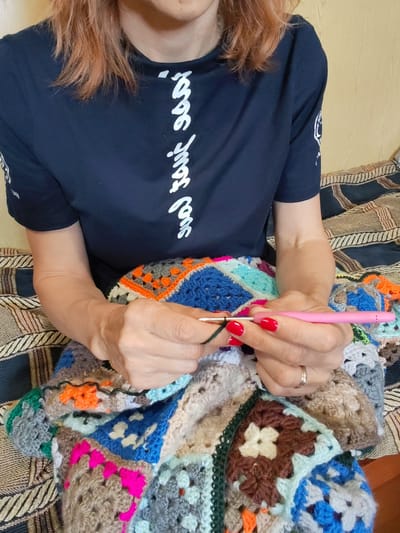 In this media file, a person is captured from the chest down, engaged in crocheting a colorful blanket. The individual, with reddish-brown hair and bright red nail polish, is wearing a dark blue t-shirt with white stylized text running vertically down the front. The t-shirt also has a white graphic on the left sleeve.
The person is seated, as indicated by the patterned upholstery of a couch or bed beneath them, which features stripes in dark blue, tan, and brown. The focus is on the hands and the crocheting process. The person holds a pink crochet hook, actively working with dark green yarn and a complex, multi-colored granny square blanket. The blanket displays a vibrant assortment of squares in shades of blue, orange, pink, grey, cream, and brown, with floral motifs visible in some of the brown squares.
The lighting suggests an indoor setting, likely during the daytime. No specific temporal or weather cues are evident, and the location context of Vytegra, Russia, is not directly discernible from visual elements within the frame. The overall mood is one of quiet concentration and creative activity. The visible text on the t-shirt is in a decorative, possibly East Asian, script.