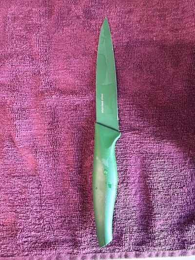 The image shows a green paring knife lying on a purple towel. The knife is oriented horizontally in the center of the image. The blade is imprinted with the phrase "stainless steel". There is no evidence to suggest that this image was taken in Arinj, Armenia. No people, subjects, objects, actions, or interactions can be discerned other than the knife resting on the towel. The environment is limited to the towel and the knife.