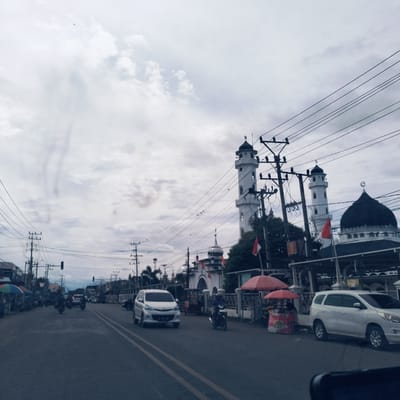 This image presents a daytime street scene in an urban or semi-urban area, identified as Barat Yaman, Indonesia. The dominant feature is a large mosque located on the right side of the street. It boasts two tall, slender white minarets, each crowned with a dark, pointed spire and a crescent moon finial. A prominent dark-domed roof also features a crescent moon, and the mosque complex is adorned with several red and white Indonesian flags.

The asphalt road, marked with double yellow lines, suggests a two-way street with ongoing traffic. A white multi-purpose vehicle (MPV) is visible in the foreground on the right, while other cars and numerous motorcycles, some with riders, populate the road further down.

Along the right curb, two street vendor stalls with vibrant red umbrellas are set up. Visible text on one stall indicates "STRAWBERRY" and a price "Rp. 10.000". Overhead, a dense web of black power lines stretches across the frame, supported by multiple utility poles that line both sides of the street.

The sky is heavily overcast with a uniform cover of white and grey clouds. A small bird can be seen flying in the upper left portion of the sky. The scene depicts a typical busy day in an Indonesian locality, characterized by local architecture, street commerce, and active transportation.