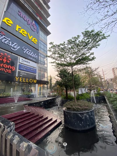 The image displays an urban scene in Narayanganj, Bangladesh, focusing on a modern commercial complex and its immediate surroundings. A multi-story building, clad in dark panels and extensive glass, occupies the left side of the frame. Illuminated signs for various establishments are visible on its facade, including "এ্যাডভান্সড" (Advanced), "e reve", "The Tasty Table", "TAAGA", "TAAGA MAN", and "YELLOW". Through the ground-level windows of the "YELLOW" store, two human figures are faintly discernible inside.

In the foreground and midground, a landscaped area features a multi-tiered water installation constructed with dark, possibly black, polished stone. Several small water fountains emit jets upwards from the water's surface. Dark reddish-brown steps, appearing to be part of a staircase, descend towards or are partially submerged within the water. Centrally located within the water feature is a circular planter, made of the same dark stone, containing a small tree with green foliage and low-lying plants. Small lights are visible embedded in the water.

To the right, beyond the water feature and scattered trees, a street or pathway is visible, populated by several indistinct individuals walking or standing. Overhead, a dense network of utility wires traverses the sky. The sky exhibits a gradient from pale blue to a soft orange-pink hue, indicative of late afternoon or early evening lighting.