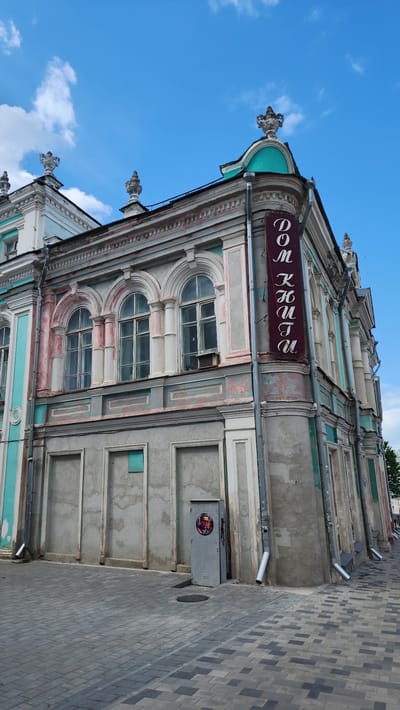 The image shows the facade of a building in Smolensk, Russia, specifically the Dom Klubova (Klubov's House). The building is ornate, with a mix of pink, turquoise, and grey paint. There are three levels of arched windows, and a maroon sign in Cyrillic script hangs on the building. A drainpipe runs down the side of the building, and an electrical box with a circular sticker is visible near the ground. The sidewalk is paved with grey and blue rectangular stones. The sky is blue with scattered white clouds.