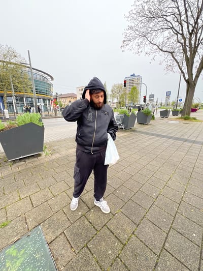 A man with a beard and wearing a black hooded jacket, black pants, and white sneakers stands on a paved sidewalk in Kehl, Germany. The man has his hands to his ears and is holding a white plastic bag in his left hand. The sidewalk is made of grey rectangular pavers and some grass grows between them. In the background, a modern, curved building with glass windows, a tall apartment building, traffic lights, street signs, and planters can be seen. A tree with bare branches is visible in the upper left corner. The scene appears to be outdoors on a cloudy day.