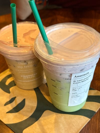 Two iced beverages with green straws are on a table, one in front of the other. The beverage in the foreground is in a clear plastic cup with a white label that reads "Anastasia Gr Iced Lavender Cream Matcha". The other beverage is in a similar cup and has a sticker on the side that says "That first sip feeling". The drinks are sitting on a brown and green Starbucks napkin. The background is out of focus and appears to be a wooden table. The scene is likely a Starbucks cafe in the City of Yonkers, United States of America, given the branding on the napkin and the style of the cups.
