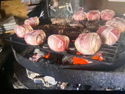 The image shows several food items wrapped in bacon and arranged on a grill over glowing embers. There are at least ten such items visible, with some appearing to be meatballs or small round portions of meat. Behind them, on the same grill, are several larger, flatter pieces of meat, likely steaks or burgers. The grill is set over a fire pit with visible red-hot coals. In the background, a person is partially visible, seemingly tending to the grill. The overall scene depicts an outdoor cooking event. The prompt mentions a location in Kerava, Finland, but there are no specific visual cues within the image to confirm this location. The background is slightly out of focus, showing wooden structures and possibly some stacked items.