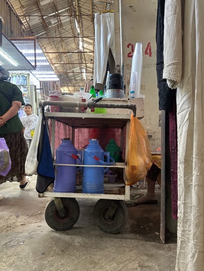 The image depicts a market scene in Kara-Suu, Kyrgyzstan. In the foreground, a utilitarian cart with two wheels is loaded with items. The cart has multiple shelves. On the middle shelf, two large insulated containers, one purple and one blue, are prominently displayed. An orange plastic bag hangs from the cart. Above these, various containers, plastic cups, and bottles are arranged on the top shelf. In the background, a young boy in a white t-shirt stands looking towards the camera. To the left, a person wearing a dark green shirt and a patterned skirt is partially visible. The environment appears to be an indoor market with a corrugated metal roof supported by metal beams. Clothing items, including white and black garments, are hanging on the right side of the image. The ground is concrete, showing signs of wear and some debris. A red number "204" is painted on the wall behind the cart.