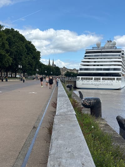 A wide-angle, eye-level shot captures a scenic waterfront promenade in Bordeaux, France, with a large cruise ship, the "Seven Seas Grandeur," docked at the quay. The promenade features a textured, light-brown paved surface with subtle markings and a grey concrete edge along the water. A wooden barrier runs parallel to the water's edge, separating the promenade from a grassy area with scattered vegetation and bollards. In the background, a line of lush green trees borders the promenade, beyond which lie buildings with architectural details, including spires. The sky is bright blue with scattered white clouds and a white contrail from an airplane. Several people are visible on the promenade; some are walking or jogging, while others are standing or sitting. The cruise ship, prominently positioned on the right side of the frame, is a large white vessel with multiple decks and windows. Two black metal bollards, one with graffiti, are visible on the quay. The water is a muted brown, consistent with a river or estuary.
