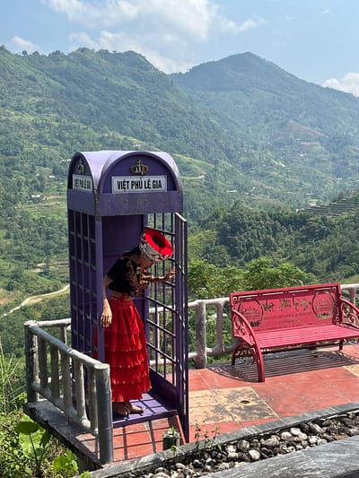 An outdoor scenic viewpoint in Thông Nguyên Commune, Vietnam, is captured on a clear, sunny day with a partly cloudy sky. The backdrop is a vast expanse of verdant mountains, some featuring visible terraced areas.

In the foreground, a person is seated inside a unique, purple cage-like structure, resembling an ornate phone booth, which serves as an observation deck or photo spot. The individual is wearing a distinctive red, pleated skirt, a dark top, and a traditional-looking red and silver headpiece. They are posed leaning against the side of the structure, gazing out at the mountainous panorama, with a relaxed demeanor.

Adjacent to the purple structure, on a platform tiled with red and dark squares, stands a decorative red metal bench with an intricate backrest. Alongside the platform, there's a section filled with small, dark rocks, and a concrete railing or staircase is visible in the lower foreground.

Visible text on the purple structure reads "VIỆT PHÚ LÊ GIA". The red bench's backrest also features text, stating "NƠI LƯU GIỮ NỀN VĂN HÓA VIỆT" (Place to preserve Vietnamese culture) and a phone number, "SDT:0948549999". The overall scene suggests a cultural or tourist attraction offering stunning views of the Vietnamese highlands.