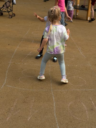 In Malakhovka, Russia, two young girls are engaged in a game of hopscotch on a tan, granular playground surface. The girl in the foreground, viewed from behind, has blonde hair in a braid and wears a tie-dye t-shirt with purple and green splotches, light blue leggings, and white shoes with pink accents. She stands with her arms outstretched, facing away from the camera and towards the hopscotch grid drawn in white and pink chalk on the ground. A hopscotch grid, with several numbered squares, is visible on the ground. Another child, partially visible in the background, wears a pink shirt and blue pants, and stands within the hopscotch grid. A stroller and wooden playground equipment are also present in the background. The lighting suggests it is daytime.