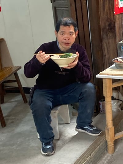 An Asian man in a black sweater and jeans sits on a wooden chair. He holds a bowl with chopsticks. He looks at the camera. The setting appears to be an indoor space with wooden walls. A wooden table sits above his head. The room's flooring is light-colored.