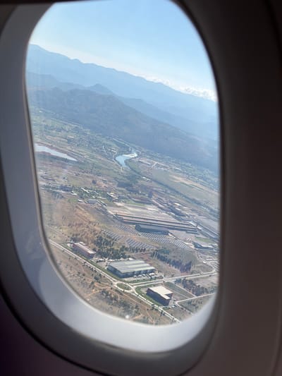 An aerial view from an airplane window shows a landscape with mountains in the distance and an industrial area below. The industrial area appears to be a large complex with multiple buildings, some with large, flat roofs, and what could be solar panels arranged in rows. A river or body of water snakes through the landscape, and roads with some vehicles are visible. The terrain is uneven, with some areas of sparse vegetation and rocky soil. The sky is clear and blue, with distant mountains fading into a hazy horizon. The image captures a broad view of the environment, focusing on the sprawling industrial development within a mountainous region.