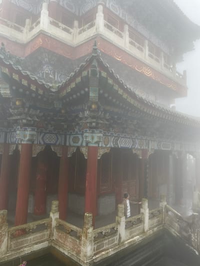 A low-angle, hazy photograph captures a section of an ornate, traditional Chinese building. The architecture features red columns supporting intricately decorated eaves in shades of blue, gold, and white. Visible through an arched opening, a person stands partially concealed, their arm reaching towards the structure. The scene is enveloped in fog, obscuring details and creating a muted color palette. The location is likely within Yongding District, China.