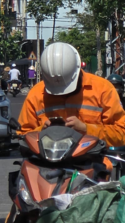 In Unknown, Vietnam, a person wearing an orange jumpsuit and a white helmet is on an orange motorcycle. The person is turned sideways, but their hand is visible on the motorcycle's handlebar. There is a green sheet or tarp covering part of the motorcycle. Other vehicles are in the background. Trees and buildings are visible behind the person and motorcycle.