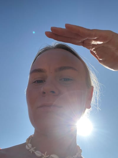 A low-angle, close-up selfie shows a person holding their hand up to shield their eyes from the bright sun. The person's face is partially illuminated, with the sun creating a lens flare effect. The sky is a clear, bright blue. The person is wearing a seashell necklace. The image appears to be taken outdoors, likely on a sunny day. There is no information in the image to suggest the location is in Ust'ye, Russia.