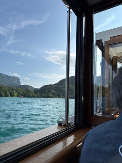 The image shows a view from inside a boat on Lake Bled in Slovenia. The boat is moving, as evidenced by the rippling water.  The camera is facing toward the front of the boat. The viewer is looking out a window, framed by wood and black trim. The window is open and shows a view of the water and surrounding mountainous terrain. The boat is in the lake, with the shoreline in the background. The sky is blue and has white clouds. The water is turquoise and choppy. There is a small island in the lake, with greenery on it. The mountainous terrain is covered in trees and foliage. The sun is shining and the weather appears calm. A person is sitting inside the boat and partially visible on the right side of the frame. The person is wearing a blue shirt and is looking out the window. The boat is in the middle of the lake. The coordinates are 46.363 latitude and 14.094 longitude. The location is Bled, Slovenia.