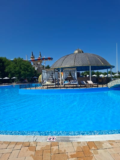 The image depicts a sunny day at a water park, likely in Ravadinovo, Bulgaria, as suggested by the location context. The scene is dominated by a large, vibrant blue swimming pool, its surface rippling under the bright sunlight. The pool is bordered by a tiled edge and a textured stone pathway. A prominent white grate runs along the edge of the pool. On the far right of the image, a small circular sign with a red border and a black silhouette of a foot with a diagonal line through it is visible, indicating a "no shoes" policy.
In the background, a large, ornate gazebo with a distinctive domed roof stands beside the pool, offering shaded seating. White curtains hang from the gazebo, suggesting a relaxation area. Beyond the gazebo, a water slide with red and blue stripes is visible, along with the turrets of what appears to be a castle-like structure, hinting at the park's theme. Lush green trees frame the background, adding to the summery atmosphere. The sky is a clear, brilliant blue, indicating fair weather and a daytime setting. No people are clearly visible, but the presence of lounge chairs and the overall setup suggest it is a recreational area where people would be engaged in swimming and leisure activities.