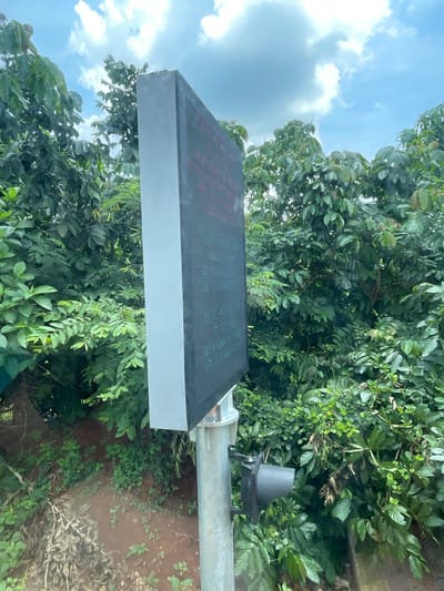 A digital information display board is mounted on a metal pole in a natural setting with lush green foliage and a cloudy sky. The display board is rectangular and black, with text visible in red and green pixels. A black mechanical device is attached to the pole below the display board. The ground is covered in soil and vegetation. The context provided mentions the location as Bình Xuân 2, Vietnam, suggesting this might be a roadside or public area within that region.