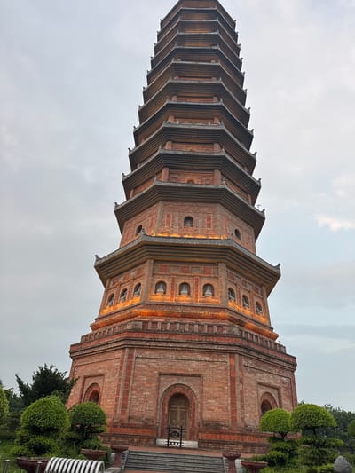This image depicts the 9-story Bai Dinh Pagoda, a prominent Buddhist temple complex located in the commune of Gia Sinh, Gia Vien district, Ninh Binh province, near the city of Tây Hoa Lư Ward, Vietnam. The pagoda is constructed of brick and features multiple tiers, each with curved, tiled roofs characteristic of traditional Vietnamese architecture. Niches containing Buddha statues are visible on several levels, illuminated by warm orange lighting. The pagoda's base is surrounded by manicured greenery, including intricately shaped bushes and trees, as well as planters and seating. The sky above is overcast, suggesting diffused daylight or evening conditions. The foreground shows a pathway leading to the pagoda entrance, with steps and decorative elements.
