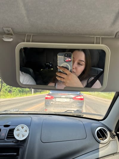 The image shows a young woman taking a selfie in a car's vanity mirror. She is wearing a light-colored tank top and has long, dark hair. She is holding a smartphone in her right hand, which is reflected in the mirror. Her left hand, adorned with a ring, is also visible. In the background of the mirror's reflection, another person wearing a light-colored t-shirt is visible in the passenger seat. The car appears to be in motion, as indicated by the road and other vehicles visible through the windshield. A dark-colored car with a license plate reading "C723BY 159" is directly in front of them. The interior of the car features a dashboard with an air freshener and a circular air vent. The scene takes place on a sunny day on a paved road, likely in a rural or suburban area of Pesterevo, Russia, given the presence of greenery and trees along the roadside.