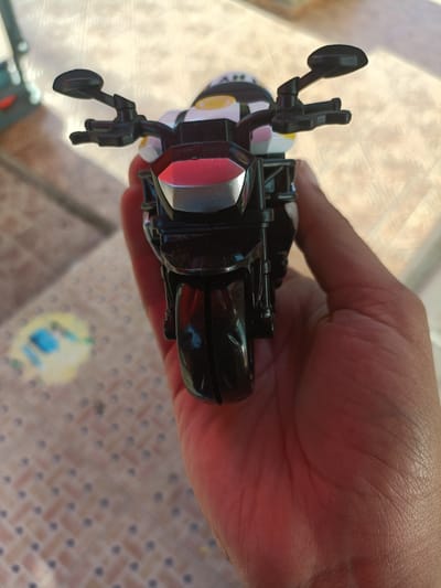 A close-up, top-down shot shows a hand holding a black toy motorcycle. The motorcycle has a red and silver front, and yellow and white accents on the helmet of the toy rider. The background is blurred, showing a tiled surface with a colorful, out-of-focus object on it. The lighting suggests an outdoor setting, possibly in the city of Safi, Morocco, as the tiled floor and warm sunlight are common elements of outdoor spaces there. The image focuses on the toy motorcycle and the hand holding it, with the environment serving as a backdrop.