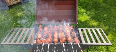 The image captures an outdoor barbecue scene, likely in a rural setting such as Пацейкаўскі сельскі Савет, Belarus, given the lush green grass and natural light. A metal barbecue grill, a common feature for outdoor cooking, is centrally placed. The grill is filled with hot coals, emitting visible plumes of smoke that rise and obscure parts of the food being cooked.
Several skewers, each laden with pieces of meat, are arranged horizontally across the grill, undergoing the cooking process. The meat appears to be seasoned and is in various stages of browning, some pieces showing char marks indicative of grilling. The skewers themselves are made of metal, with spiral-shaped handles at the visible ends, suggesting they are designed for easy turning.
The grill has a hinged lid, which is open and angled upwards, allowing the smoke to escape. The grill is supported by a dark-colored metal frame, and to its left, there's a slatted metal rack extending outwards, possibly for placing food or utensils.
The surrounding environment is dominated by a vibrant green lawn, dotted with small white flowers, consistent with a natural outdoor space. In the upper left corner of the image, a portion of a colorful textile is visible, possibly a tablecloth or a decorative fabric, hinting at a gathering or picnic. The overall impression is one of a casual, enjoyable outdoor meal preparation.