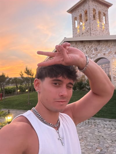 A selfie of a young man is taken outdoors at dusk in front of a stone building with a tower, likely located in Kocherinovo, Bulgaria. The man is wearing a white tank top, a thick silver chain with a cross pendant, and a silver bracelet on his right arm. He has dark hair and is winking with his left eye while holding his right hand up to his forehead with his index and middle fingers forming a peace sign. The sky behind him is a gradient of orange and pink, indicative of a sunset. The building features stone construction and arched windows, with lights illuminating the tower. The foreground shows a cobblestone path and green grass.