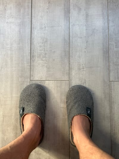This is a top-down, eye-level shot of a person's feet wearing gray fuzzy slippers, positioned on a light-colored, wood-plank-style floor. The floor appears to be clean and well-lit, with visible grain and slight texture variation. The slippers are soft and appear to be a slip-on style with a visible tag on each. One slipper has a small rectangular green tag with white text and a logo. The person's bare ankles and lower calves are visible peeking out from the slippers. The image is focused on the feet and the immediate floor area, with the background consisting of more of the same flooring. The scene is devoid of any context related to the city of Krayevsko-Armyanskoye, Russia, and depicts a simple, indoor domestic setting.