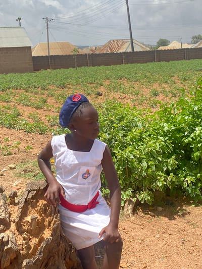 A person wearing a white uniform, red sash, and navy blue cap with the letters "G. B." visible on the cap lies on a weathered tree trunk in Turu, Nigeria. The individual is positioned in a field with sparse vegetation. In the background, there are buildings with corrugated metal roofs and block walls under a cloudy sky. A patch of dense green foliage is located near the tree trunk. A power pole and lines are visible against the sky. The ground is dry and appears to be dirt or sand.