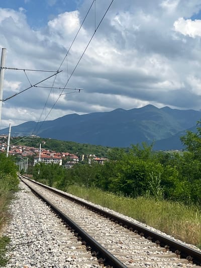 The image, when viewed horizontally, captures a picturesque landscape featuring a railway line, a town, and large mountains under a partly cloudy sky. In the foreground, two parallel train tracks are prominently visible, laid on a bed of light-colored gravel ballast. Green grass and various leafy plants line the immediate vicinity of the tracks on the right side.
Spanning the upper left portion of the sky, a complex network of overhead electrical lines (catenary system) is suspended, indicating an electrified railway. Beyond the tracks and the surrounding greenery, a town or village is situated, with numerous buildings featuring characteristic orange or reddish-brown tiled roofs. This settlement lies at the base of a series of large, dark green mountains that form the dominant background. The mountains appear rugged and forested, with their peaks partially obscured by a slight atmospheric haze.
The sky above is a mix of bright blue and large, fluffy white and grey clouds, suggesting a clear, bright day, likely late morning or early afternoon. No people or specific activities are depicted in the scene, which primarily focuses on the landscape and infrastructure. There is no visible text. The combination of railway, mountainous terrain, and clustered housing is consistent with the provided context of Dupnitsa, Bulgaria.