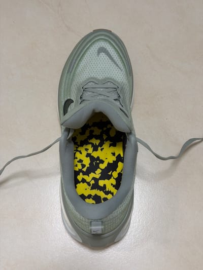 A top-down, medium shot depicts a light green Nike running shoe with a yellow and black patterned insole resting on a beige tiled surface. The shoe's laces are untied. The location is unknown and cannot be determined from the image. There is no identifiable information to suggest Kfar Saba, Israel, as the location. There are no people visible.