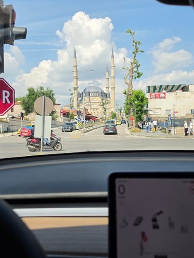 The image captures a street scene in Edirne Merkez, Turkey, observed from the interior of a vehicle, looking towards the prominent Selimiye Mosque under a bright blue sky with large white cumulus clouds and faint contrails.

In the bottom foreground, the edge of a dark steering wheel is visible on the left, and a blurred digital display, likely a vehicle's central screen, is in the bottom right, showing "0" and various icons, including a red stop light representation.

The midground features an asphalt road with white and yellow markings. A red delivery scooter, emblazoned with "Bizde her yer Domino's Pizza" on its rear box, is positioned center-left, with the rider visible. Behind it, a yellow taxi is partially discernible. Further down the road, a dark grey sedan is turning left. Traffic infrastructure includes a red octagonal stop sign with a white "R" on the far left and a black traffic light pole. On the right, a building with a red "ELARA CAFE" sign and a green striped awning is visible, accompanied by smaller Turkish text signs. A tall, somewhat bare tree with new green leaves stands prominently on the right side of the street. Multiple pedestrians are seen walking on the sidewalks on both sides, with a group of approximately five individuals on the right sidewalk. Iron railings line the sidewalk on the right.

The background is dominated by the Selimiye Mosque, an iconic Ottoman imperial mosque with a large central dome, numerous smaller domes, and four tall, slender minarets culminating in pointed spires.