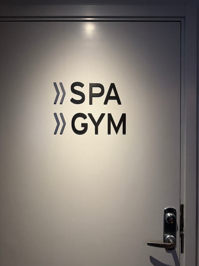 A close-up, eye-level shot shows a metal door in what appears to be a hotel or facility hallway in Östersund, Sweden. The door is painted a light gray and has a dark gray frame. Two sets of dark gray, raised lettering are affixed to the door, indicating "SPA" on the upper line and "GYM" on the lower line. Each word is preceded by a decorative symbol resembling two superimposed arrows pointing to the right. Below the lettering, a brushed metal door handle and lock mechanism are visible. The lighting casts a shadow from the handle onto the door. The background shows the edge of the door frame and a hint of the surrounding wall.