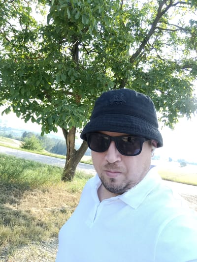 A man wearing a black bucket hat, sunglasses, and a white polo shirt is taking a selfie outdoors. He has a goatee and is wearing a wireless earbud in his right ear. Behind him, a tree with green leaves stands in a grassy area. A paved road is visible to the left, and a hazy landscape with fields and trees extends into the background. The image appears to be taken in the city of Šternov, Czechia, based on the metadata.