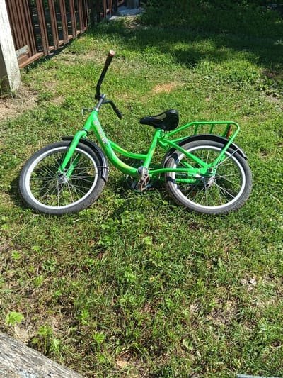 A bright green bicycle is lying on its side on a grassy lawn in Ostrovtsy, Russia. The bicycle is a Stels brand, with a basket on the back and white-rimmed wheels. The handlebars are black and the seat is black with a patterned texture. The bicycle appears to be in good condition. The grass is green and well-maintained, with some patches of dry earth visible. In the background, there is a wooden fence and a concrete structure. The sun is shining, casting shadows on the grass.