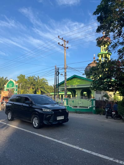 The image captures a street scene in Bukitpanjang Dua, Indonesia, bathed in the warm light of what appears to be late afternoon. The sky is a vibrant blue, streaked with wispy white clouds, suggesting clear weather. Power lines crisscross the frame, suspended from a utility pole, hinting at the presence of infrastructure in the area.

Dominating the left side of the image is a mosque, its striking green structure punctuated by a dome and a tall, slender minaret. The mosque is partially obscured by the lush green foliage of trees, adding a natural element to the architectural scene. In front of the mosque, a decorative metal gate is visible, suggesting an entrance.

A black car, a Toyota with a license plate reading "BK 1997 AEK," is parked on the right side of the asphalt road, which is marked with a white line. The car is angled, revealing its side profile. A motorcycle is also visible near the mosque, parked alongside a fence. The overall scene conveys a sense of a tranquil, everyday moment in a community that blends religious and residential elements with essential services like electricity. There is no discernible activity or event taking place beyond the static presence of vehicles and buildings.