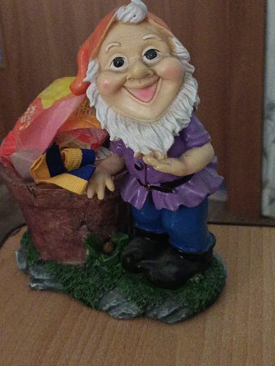 This media file features a garden gnome figurine. The gnome has a rosy-cheeked, smiling face with a white beard and eyebrows, and wears a red hat, a purple tunic, and blue pants. The gnome is leaning against a basket filled with colorful items, including what appears to be fabric scraps in shades of pink, yellow, and blue, resembling a small pair of socks. The figurine is placed on a green, grassy base with gray rocks, suggesting an outdoor or garden setting, though it is displayed indoors on a wooden surface. The background is a plain brown wall, possibly wood-paneled. The lighting is warm, and there are no indications of the time of day or weather. The overall mood of the figurine is cheerful and whimsical. No visible text is present in the image.