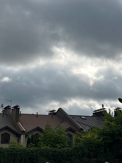 The image shows the rooftops of several houses with dark grey, cloudy skies above. The houses have tiled roofs, some with chimneys, satellite dishes, and antennas. Trees and dense greenery are visible in front of the houses. The scene appears to be a residential area in Yurlovo, Russia.