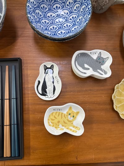 The image shows a collection of ceramic dishes and chopsticks laid out on a wooden surface. In the top left is a stack of blue and white patterned ceramic bowls. To the right of the bowls are three small, irregularly shaped white ceramic dishes, each with a different cat illustration and the word "MEOW!". The top dish depicts a grey cat, the middle dish shows a black and white cat, and the bottom dish features a ginger cat. To the left of the cat dishes is a black box containing a set of chopsticks, with two pairs visible: one pair appears to be a light brown wood, and the other pair is a dark teal color. On the far right of the image, a portion of a yellow, textured ceramic dish is visible. The overall arrangement suggests a display of tableware or decorative items. There are no people or actions depicted. The environment appears to be indoors, with the wooden surface suggesting a table or countertop. The image was taken in the city of Bellingen, Australia.