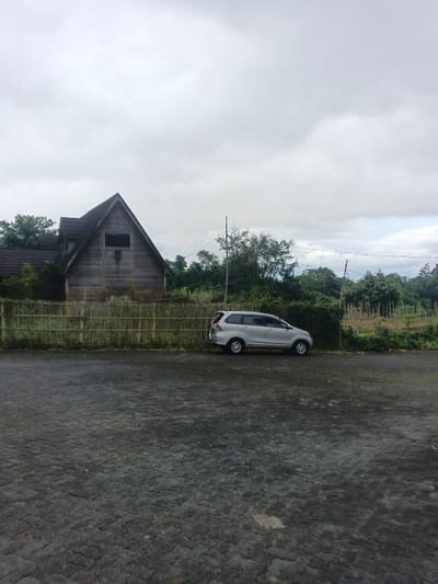 The image displays an outdoor scene in Masaiti, Zambia, under an overcast sky. The foreground consists of a dark grey, textured ground, possibly pavement or packed earth. A light brown wooden or bamboo fence spans horizontally across the mid-ground. To the left, behind the fence, stands a two-story building constructed of light brown wooden siding, featuring a steep, dark gable roof and a small, dark rectangular opening on the upper gable. A silver minivan is parked parallel to the fence, positioned behind it and facing right. Behind the fence and vehicle, dense green foliage, including trees and bushes, is visible, with cultivated land further to the right. A utility pole with horizontal wires is present behind the fence, right of the building. No individuals are visible.