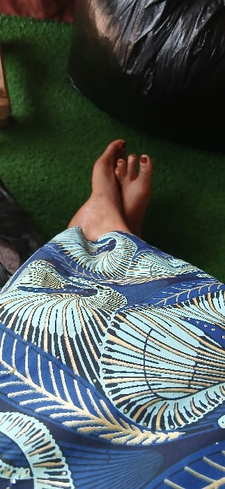 The scene depicts the lower extremities of an individual in Sabon Gari South, Nigeria. The subject's feet are crossed at the ankles, resting on a surface covered with green artificial turf. The skin tone of the feet is dark, and toenails are painted with a dark red polish. A fabric garment, possibly a skirt or dress, with a blue, light blue, and gold geometric pattern, covers the lower lap and upper legs. In the upper right background, a large, dark, crumpled object, consistent with a black plastic bag or tarp, is visible. A small section of a light brown object is present in the upper left background. No other individuals or interactions are discernible.