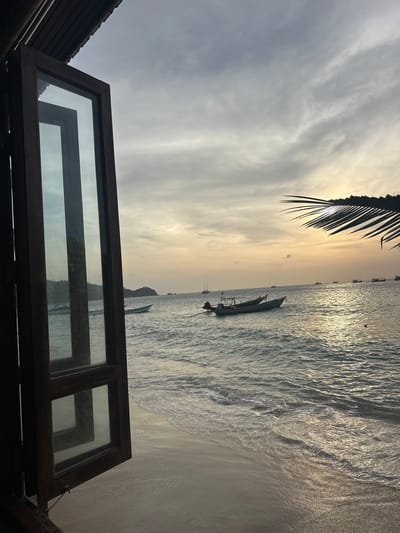 The image displays a coastal scene in Koh Tao, Thailand, observed through an open, dark-stained wooden window frame. The frame, positioned on the left, features two glass panes, each divided horizontally by a wooden crossbar. Above the window, a dark wooden ceiling structure is visible.
In the foreground on the right, a silhouetted palm frond extends diagonally from the upper right, partially obscuring the sky. The midground features a sandy shoreline at the bottom, marked by small waves breaking and receding. The ocean surface exhibits ripples and waves, reflecting ambient light.
Numerous boats are present on the water. Immediately outside the window frame, a light-colored boat is partially visible. To the right of the window, a dark longtail boat with an outboard motor is anchored. Further out, a collection of additional longtail boats and two sailboats with visible masts are distributed across the water. A small, rocky island or headland is visible in the distant left.
The sky is overcast, displaying a gradient from grey clouds overhead to warmer golden-orange hues near the horizon, indicative of low sun angle. No individuals are visible in the scene.