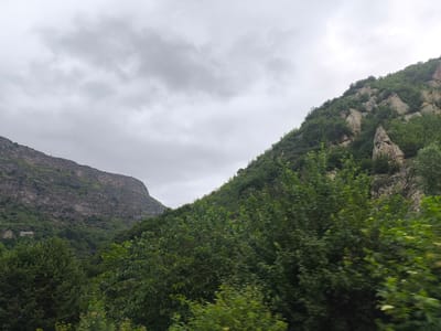 The image depicts a lush, mountainous landscape under a cloudy sky. Two prominent mountains dominate the scene, one on the left with a rugged, terraced appearance covered in sparse vegetation, and another on the right, more densely forested with trees clinging to its rocky slopes. The foreground is filled with a thick canopy of green trees, suggesting a forest or dense woodland. The overall impression is one of natural, untamed beauty.

The time of day is difficult to ascertain precisely due to the overcast conditions, but the diffused light suggests it could be late morning or afternoon. The weather appears to be cloudy, potentially indicating an approaching rain or simply a persistent overcast day.

A small, stone building is visible nestled into the side of the left mountain, adding a touch of human presence to the otherwise wild scenery. The building seems to be of traditional architecture, possibly an old house or a historical structure. Its presence hints at a settlement or a remote dwelling within this mountainous region.

The location cues, combined with the provided context of Tumanyan, Armenia, suggest a mountainous area known for its natural landscapes and potentially historical sites. There is no visible text in the image. The overall mood conveyed by the image is one of tranquility and the vastness of nature.