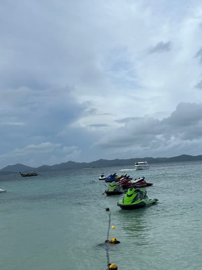 The image depicts a tranquil scene of the ocean near Tambon Phru Nai, Thailand. The water is a clear turquoise, with visible buoys and a rope marking a boundary. Several jet skis are moored in the water, with three in the foreground and several more further back. In the distance, a longtail boat navigates the water, with a speedboat approaching from the right. The horizon is lined with lush green hills, shrouded in a light mist under a cloudy sky. The overall atmosphere is serene and peaceful, conveying a sense of a quiet day at sea.