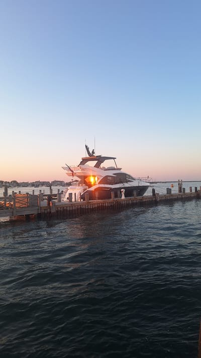 A large white and black yacht is docked at a wooden pier. The yacht has multiple levels and a dark roof. The sun is setting, casting an orange glow on the yacht and the water. The sky is a gradient of blue and pink. In the background, there are houses and a few other boats. The water is calm with ripples. This scene is likely in Nantucket, Massachusetts, a popular destination for yachts and coastal living.