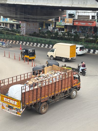 The image captures a street scene in Shamshabad, India, under an elevated overpass. In the foreground, a large, rusted brown truck filled with garbage is parked or moving slowly. The truck's rear displays the "EICHER" brand and an "AP31TU 9509" license plate, along with a "STOP" sign. Beside the truck, a large white tarp-covered object is visible. Behind the truck, a yellow and black auto-rickshaw is stopped, and a black auto-rickshaw is also present. Further back, a white panel van and a white SUV are on the road. On the right side of the frame, two individuals on a motorcycle are riding away from the viewer. In the background, buildings line the street, with various storefronts and signage, including "happi" and "Coca-Cola." A green fence separates the road from the sidewalk area. The overall environment appears to be an urban street with moderate traffic.